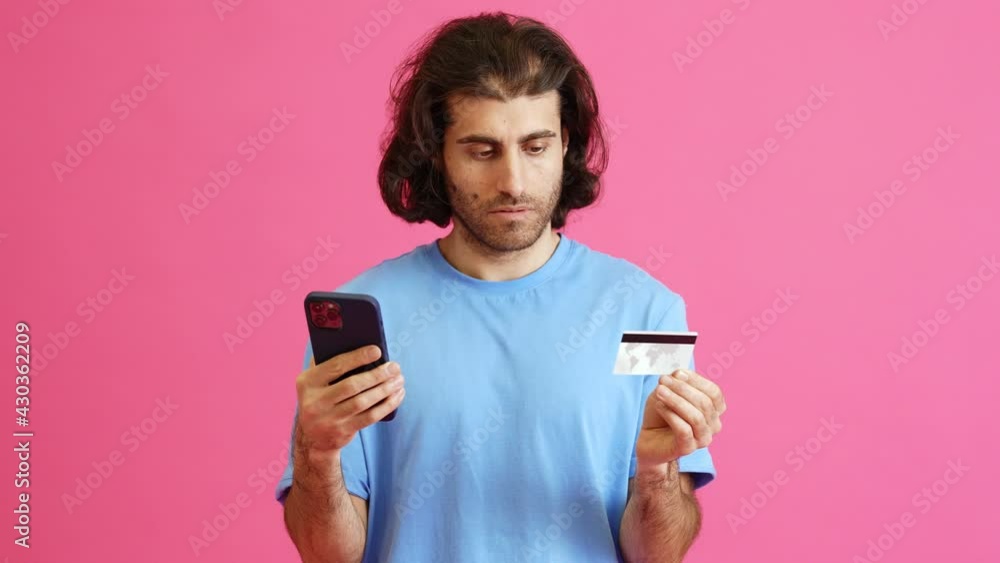 The angry man with long hair entering a card number and holding a phone in the hand while standing in a pink studio The angry man with long hair entering a card number and holding a phone in the hand while standing in a pink studio