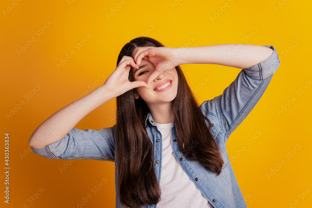 Closeup portrait of woman fingers show heart gesture look watch cover eyes isolated yellow wall background Closeup portrait of woman fingers show heart gesture look watch cover eyes isolated yellow wall background