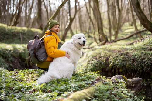 Young woman in hiking clothes and backpack sitting near big white dog in green spring forest. Enjoys and explore of tranquil nature.