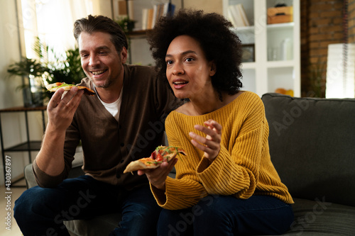 Cheerful young couple sitting on sofa at home. Happy woman and man eating pizza while watching a movie..