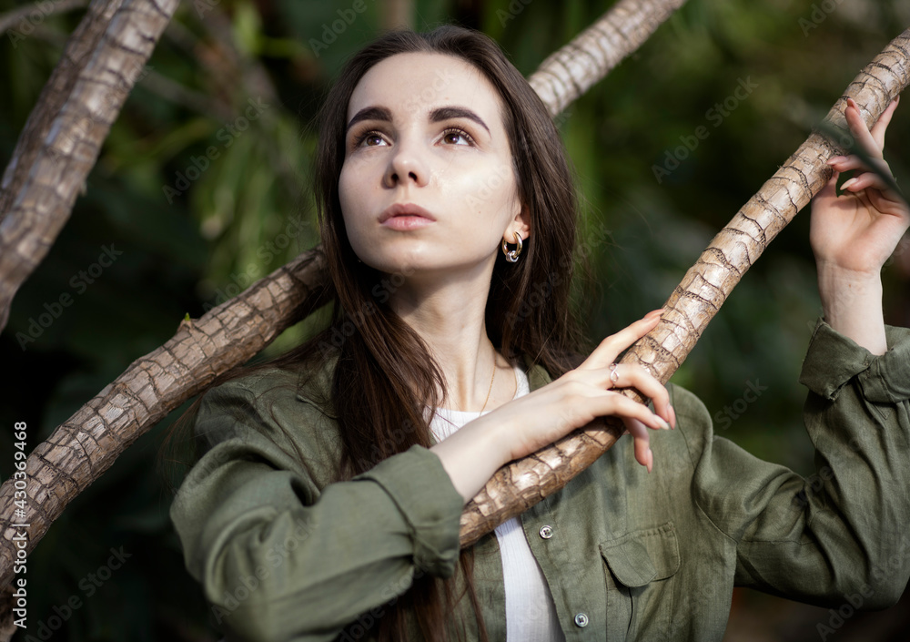 A fair-skinned girl with dark long hair, earrings, in a cotton shirt, stands near a tree, among the branches and looks up. Problems of traveling abroad for Russians, travel 2021, loneliness, earth day
