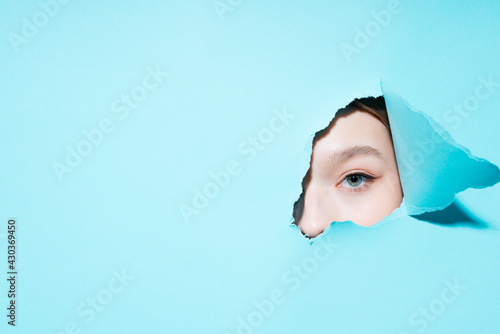 Photo of a painted eye of a girl looking at the camera through a hole on a blue background © DmitryStock