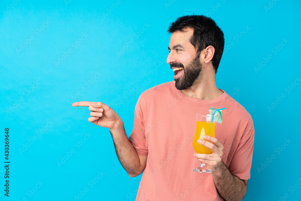 Young man over holding a cocktail over isolated blue background pointing finger to the side and presenting a product Young man over holding a cocktail over isolated blue background pointing finger to the side and presenting a product