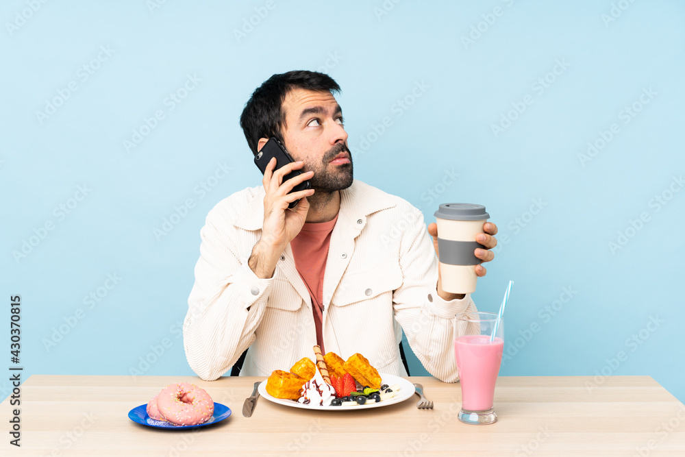 Man at a table having breakfast waffles and a milkshake holding coffee to take away and a mobile Man at a table having breakfast waffles and a milkshake holding coffee to take away and a mobile