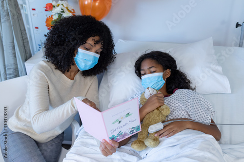 Mixed race mother and sick daughter in face masks in hospital, woman holding card and girl teddy