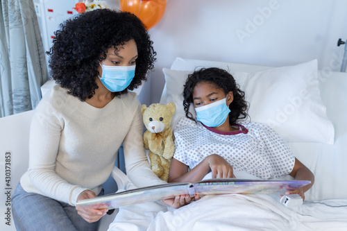 Mixed race mother and sick daughter in face masks in hospital, reading book with teddy bear