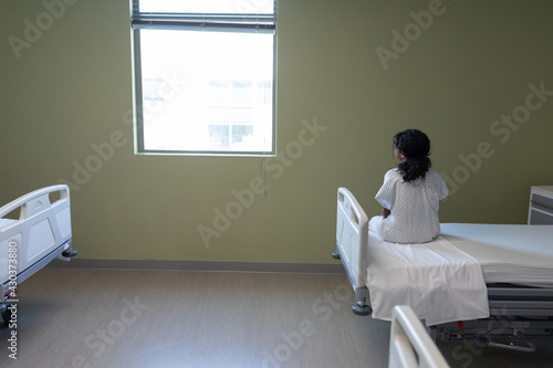 Rear view of sick mixed race girl sitting on hospital bed looking out of window