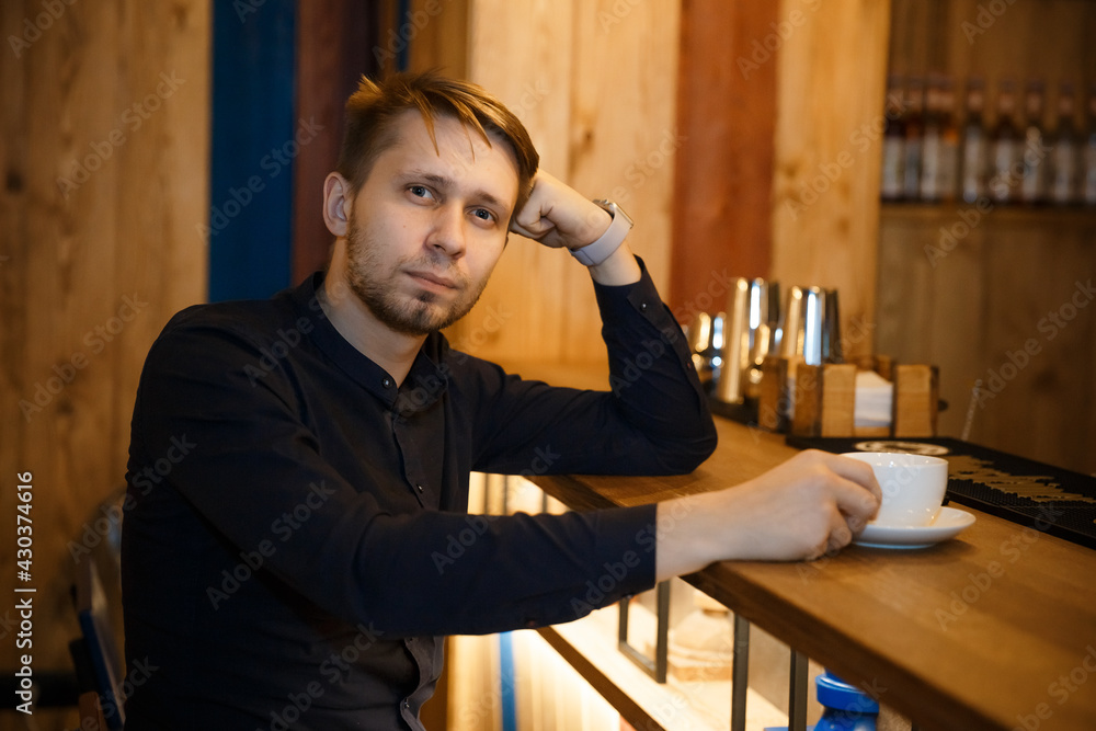 Obraz premium Close-up portrait of a young man sitting at a bar counter in a cafe with his head resting on his hand. Holds a cup of coffee and looks at the camera