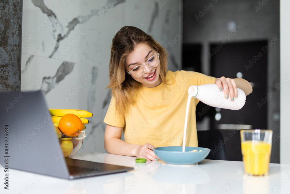 Pretty young woman having breakfast while sitting at the kitchen table, working on laptop computer Pretty young woman having breakfast while sitting at the kitchen table, working on laptop computer