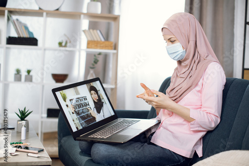 Side view of young Muslim woman sitting on sofa at home in protective mask and using laptop pc while talking with confident experienced Arabian doctor online about her symptoms and throat pain.