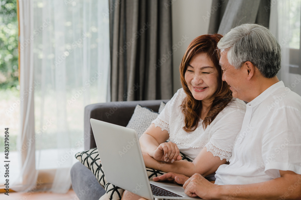 Asian senior couple video call at home. Asian Senior Chinese grandparents, using laptop video call talking with family grandchild kids while lying on sofa in living room at home concept. Asian senior couple video call at home. Asian Senior Chinese grandparents, using laptop video call talking with family grandchild kids while lying on sofa in living room at home concept.