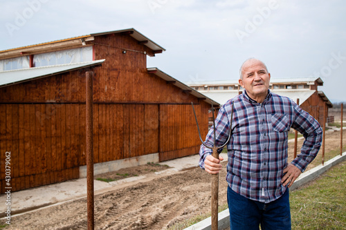 Portrait of successful farmer with pitchfork tool. In background wooden farmhouse.