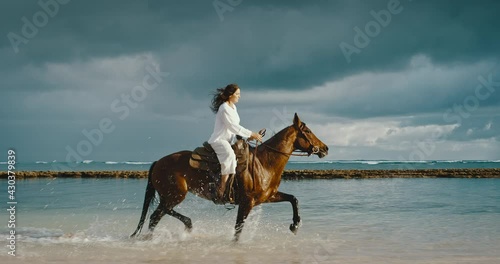 Beautiful young woman horseback riding on the beach, strong and powerful horse splashing in the water, cinematic slow motion