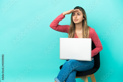 Young caucasian woman sitting on a chair with her pc isolated on blue background looking far away with hand to look something © luismolinero