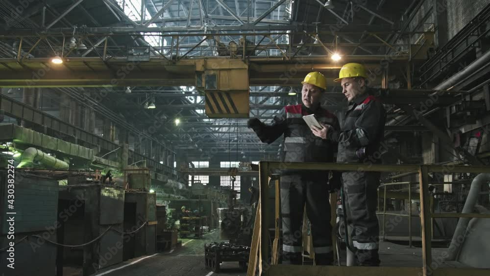 Full shot of two male tractor plant workers with digital tablet standing at foundry workshop having conversation during shift pointing at machine equipment Full shot of two male tractor plant workers with digital tablet standing at foundry workshop having conversation during shift pointing at machine equipment