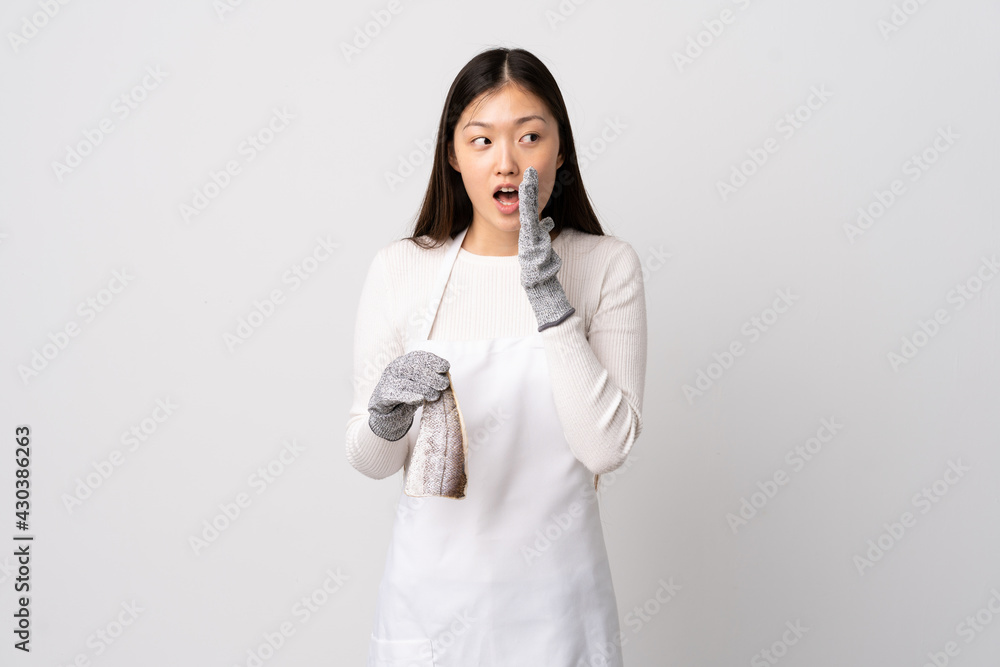 Chinese fishmonger wearing an apron and holding a raw fish over isolated white background whispering something with surprise gesture while looking to the side Chinese fishmonger wearing an apron and holding a raw fish over isolated white background whispering something with surprise gesture while looking to the side