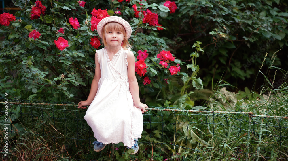 Little beautiful cute blonde girl in a gentle pink dress and hat near the bushes of a rose hip tea rose. Summer kid portrait, A funny girl sits on a hedge under rose bushes