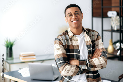 Portrait of a handsome successful confident young mixed race latino man with glasses, stylishly dressed, standing near work desk with arms crossed, looking at camera with friendly smile photo