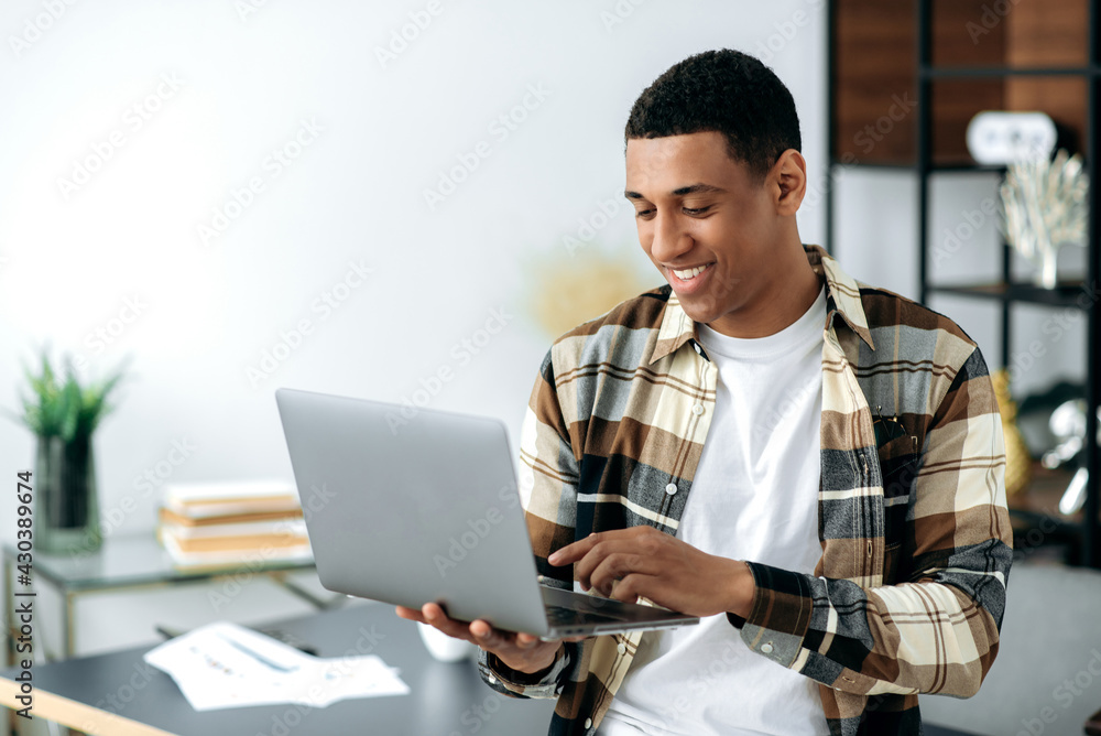 Smiling modern stylishly dressed mixed race latino freelancer guy, manager or student, stands near work desk, holds laptop, chatting online with friends, browses social networks, internet,checks email Smiling modern stylishly dressed mixed race latino freelancer guy, manager or student, stands near work desk, holds laptop, chatting online with friends, browses social networks, internet,checks email