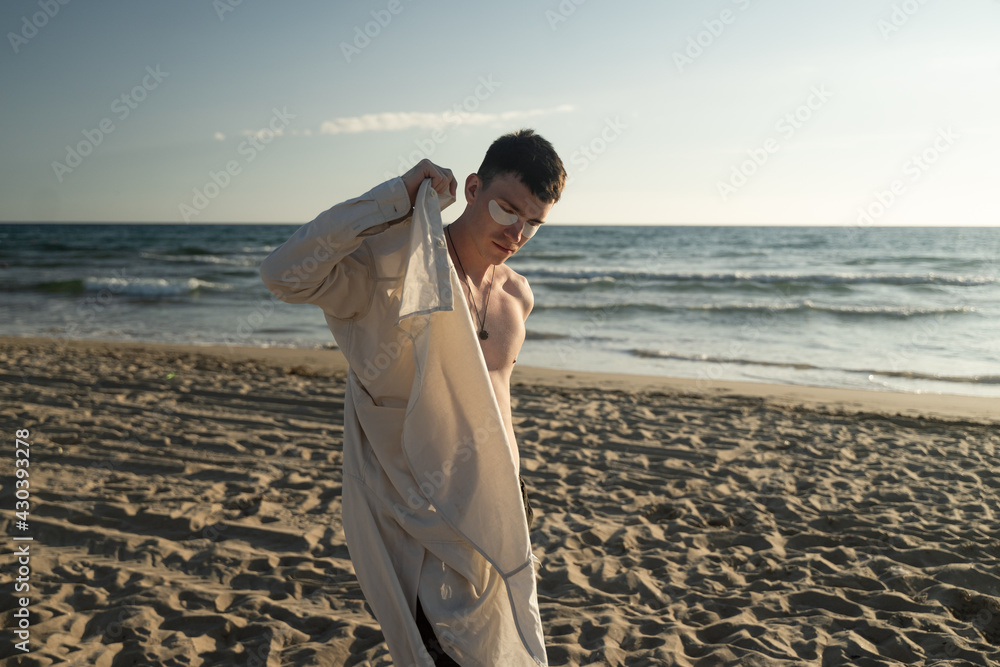 a young guy puts on a beige shirt by the sea with white patches on his face