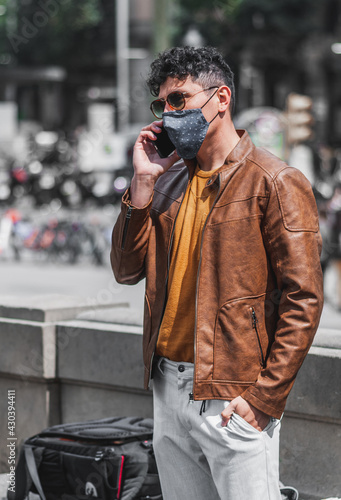 Good looking young adult man wearing casual spring clothes outdoor, talking and texting on phone in padendemic day taking measure with face mask.