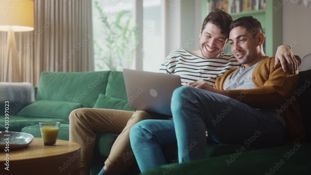 Portrait of Gentle Gay Couple Using Laptop Computer, while Sitting on a Couch in Cozy Stylish Apartment. Adult Boyfriends Online Shopping on Internet, Watching Funny Videos on Streaming Service.