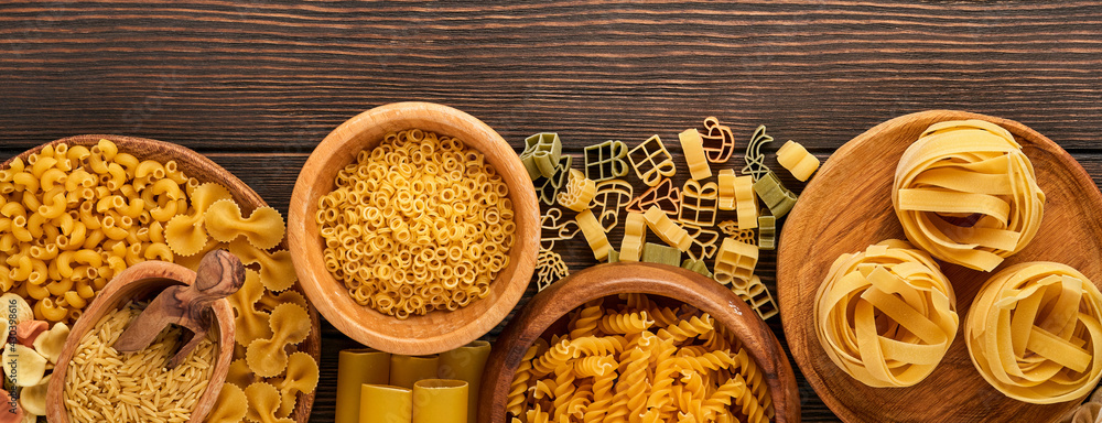 Variety of traditional italian pasta: colorful spaghetti, tagliatelle, farfalle, penne, ptititm, noodle, fusilli, cannelloni on an old wooden background. Top view with copy space. Banner.