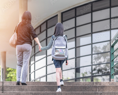 Educational back to school, parent's day, bring kid to work concept with elementary student girl child carrying backpack holding mother's hand walking up admission office building going to class