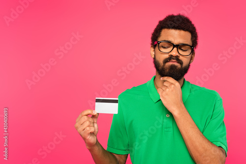 pensive african american man in glasses holding credit card isolated on pink