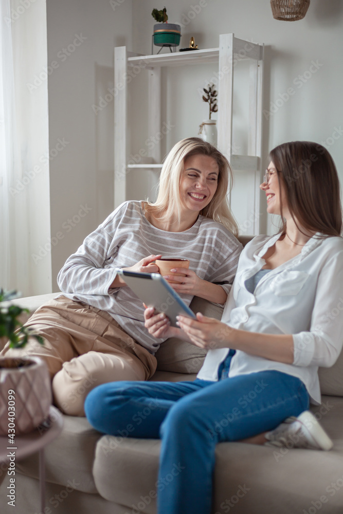 Two young women friends using tablet together, sitting on the couth