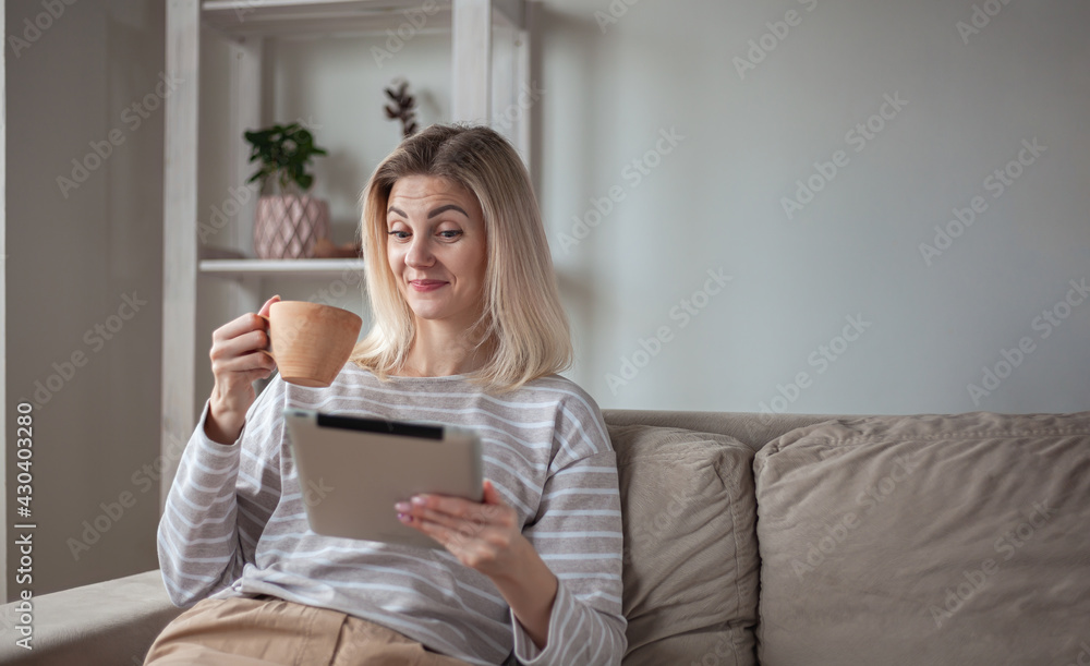 Surprised Young woman using tablet, sitting on sofa at home, drinking coffee Surprised Young woman using tablet, sitting on sofa at home, drinking coffee