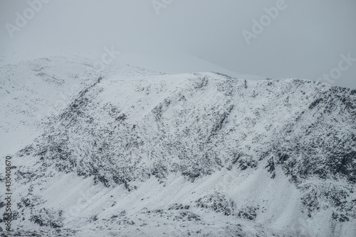 Foggy mountain landscape with white snow on black rocks in cloudy sky. Misty mountain minimalism of snowbound mountain top in low clouds. Minimalist nature background of snowy mountain peak in fog.