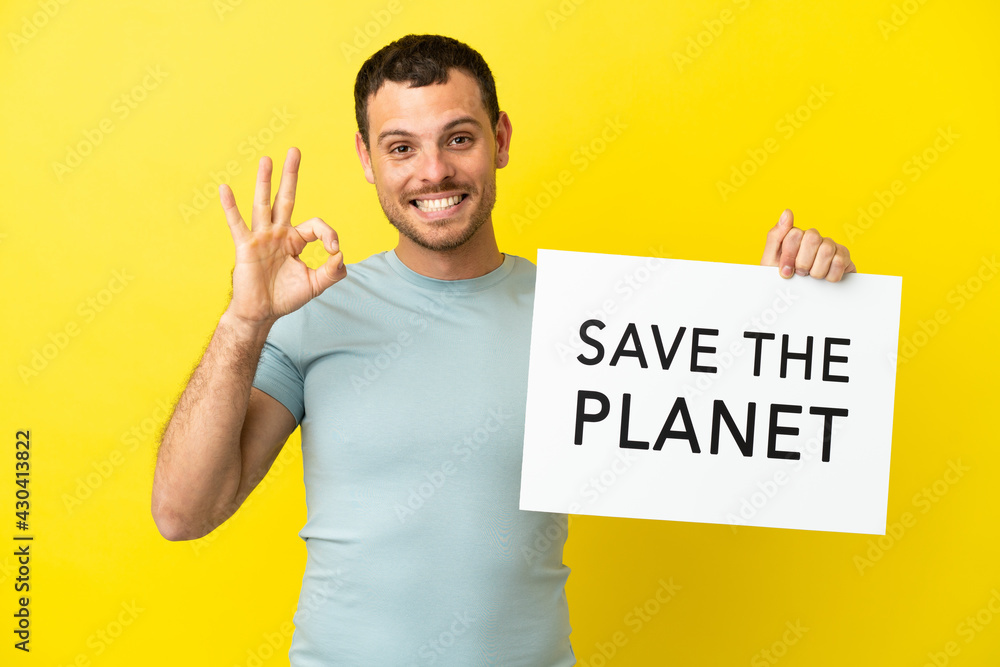 Brazilian man over isolated purple background holding a placard with text Save the Planet and celebrating a victory Brazilian man over isolated purple background holding a placard with text Save the Planet and celebrating a victory