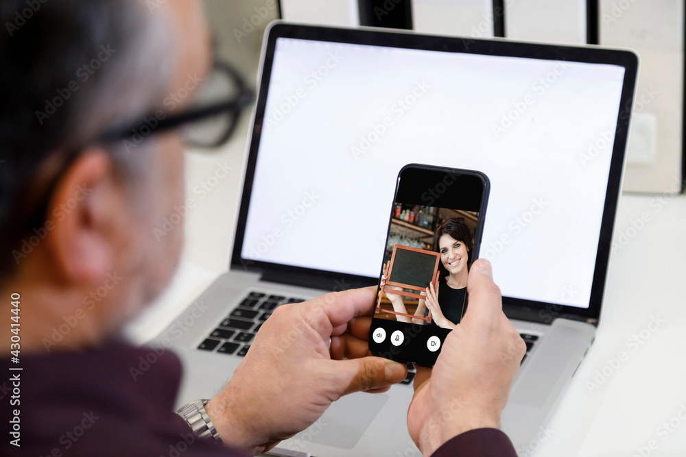 Un uomo si collega con il suo cellulare  in video conferenza con una ragazza in remoto  che gli mostra una lavagna da un locale - sullo sfondo si vede un computer con lo schermo bianco 