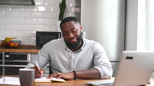 Posotive African American man sitting at the table at home in the modern kitchen, using the laptop for distance video communication, studying, working, meeting online, and making notes photo