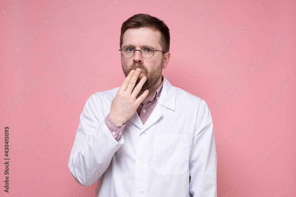 Alarmed young doctor in glasses and a white coat covered mouth with hand and looked fearfully into the camera. Pink background. Alarmed young doctor in glasses and a white coat covered mouth with hand and looked fearfully into the camera. Pink background.