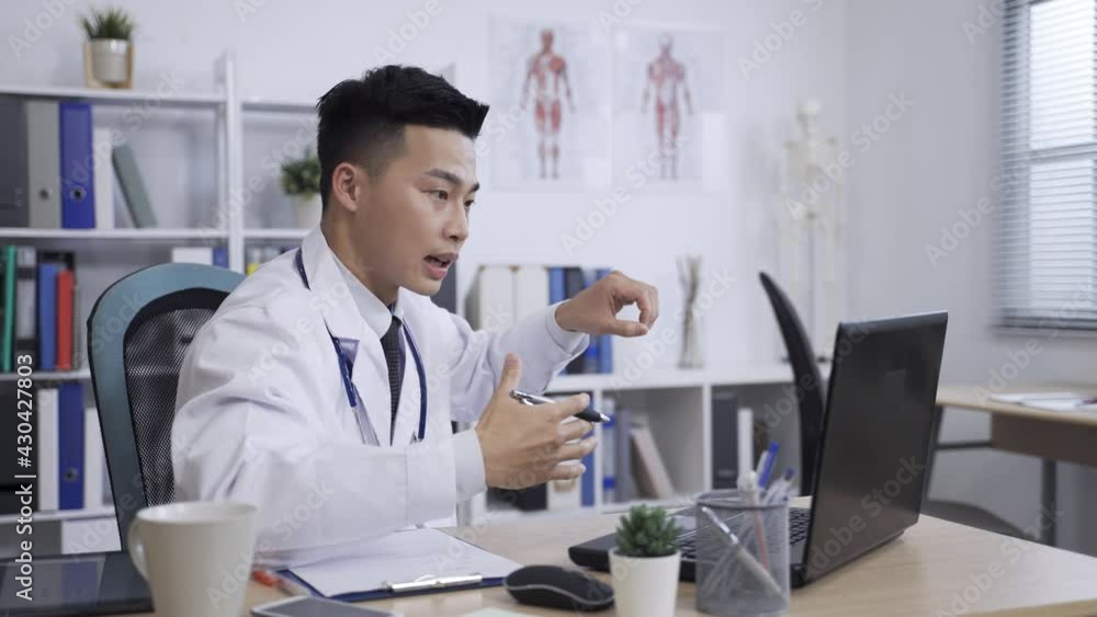asian male doctor is gesturing with a pen in hand while sharing information during an distant consultation with his patients on the computer in clinic office. asian male doctor is gesturing with a pen in hand while sharing information during an distant consultation with his patients on the computer in clinic office.