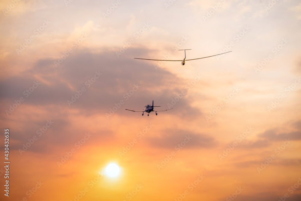 a glider sailplane towed by a small private airplane toward the evening setting sun with orange clouds