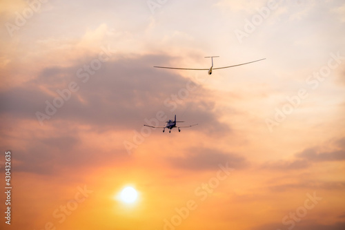 a glider sailplane towed by a small private airplane toward the evening setting sun with orange clouds