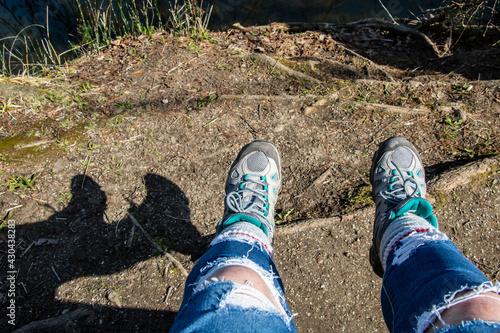 Woman's legs hanging off of a bench, feet swung forward over dirt ground. The shadow of her legs is visible, her clothing casual, ripped jeans and hiking boots.