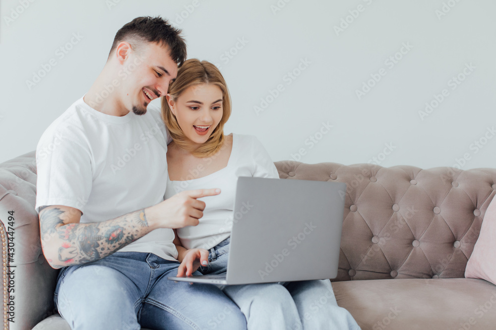 Photo of excited surprised young loving couple sitting on sofa indoors at home using laptop computer.