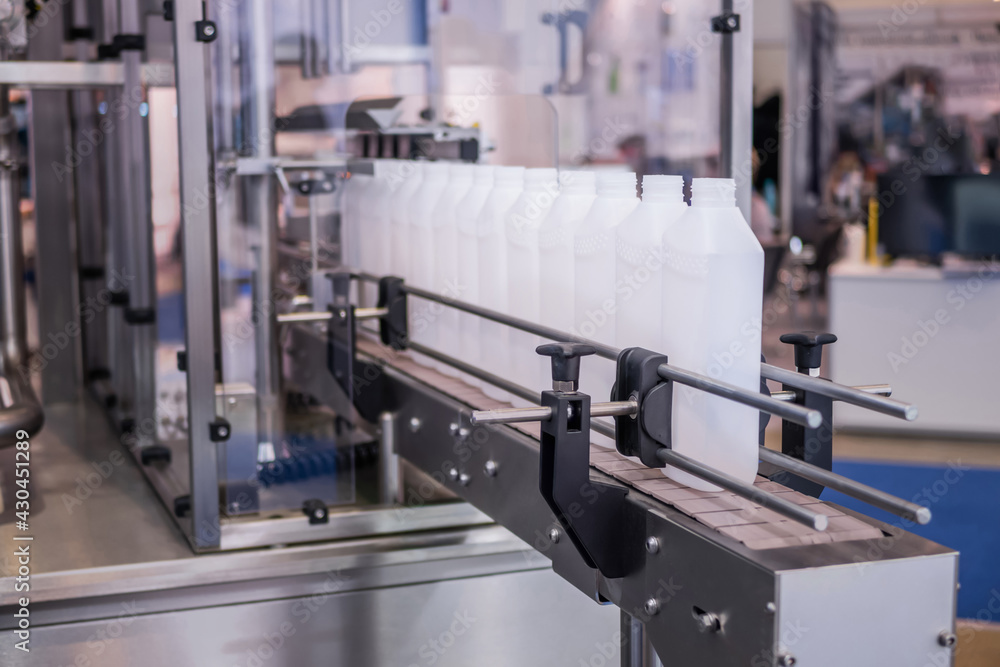 Moving polypropylene white bottles on conveyor belt of automatic liquid filling machine at plastic exhibition, trade show. Manufacturing, industry and automated technology equipment concept Moving polypropylene white bottles on conveyor belt of automatic liquid filling machine at plastic exhibition, trade show. Manufacturing, industry and automated technology equipment concept
