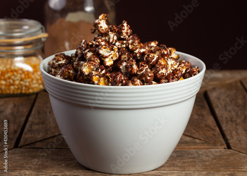 Sweet or sugared popcorn with chocolate or cocoa on wooden table and ingredients in the background.