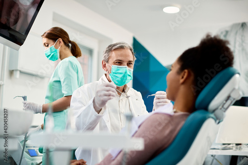 Slika na platnu Smiling dentist with face mask talking to Black woman during dental procedure at dental clinic