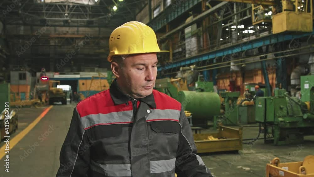 Medium shot of serious caucasian male supervisor in yellow hard hat walking along assembly shop of big tractor plant inspecting machinery equipment and work of employees Medium shot of serious caucasian male supervisor in yellow hard hat walking along assembly shop of big tractor plant inspecting machinery equipment and work of employees