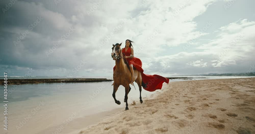 Beautiful woman horseback riding on the beach in flowing red dress, cinematic slow motion Beautiful woman horseback riding on the beach in flowing red dress, cinematic slow motion