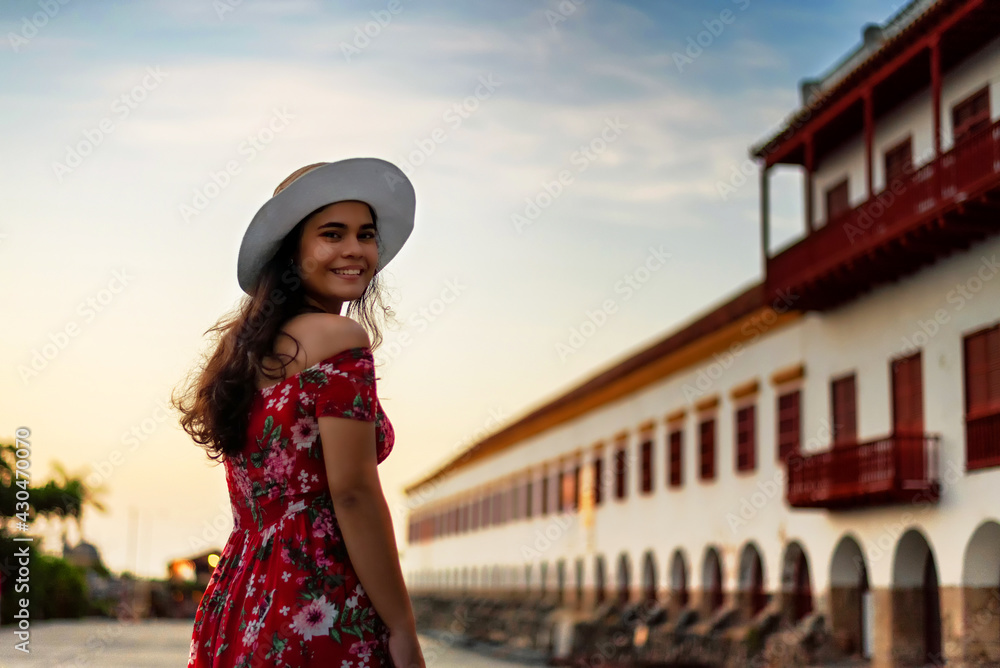 Beautiful Latina woman in red dress and white hat on a wide walkway near old building at sunset