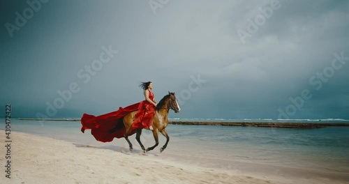 Beautiful woman horseback riding on the beach in flowing red dress, cinematic slow motion