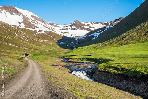 Deserted winding rough road running along a creek at the foot of snow-capped peaks in a majestic mountain scenery in Iceland on a sunny summer day photo