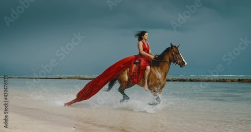 Beautiful woman horseback riding on the beach in flowing red dress, galloping in the water, cinematic slow motion